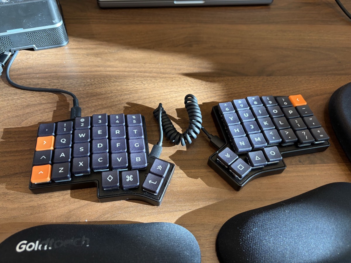Keebio Iris CE split column-stagger keyboard with dark grey keycaps and orange accent keys, connected by a coiled cable